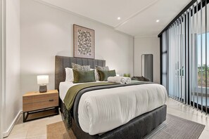 Une chambre à coucher moderne avec un grand lit, une table de chevet, une lampe de chevet, une œuvre d’art encadrée au mur et une vue sur l’extérieur grâce à une porte coulissante.