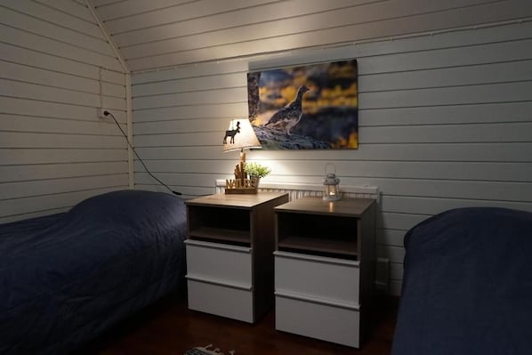 A room with two beds, a nightstand with a lamp, a painting on the wall, and a small plant.