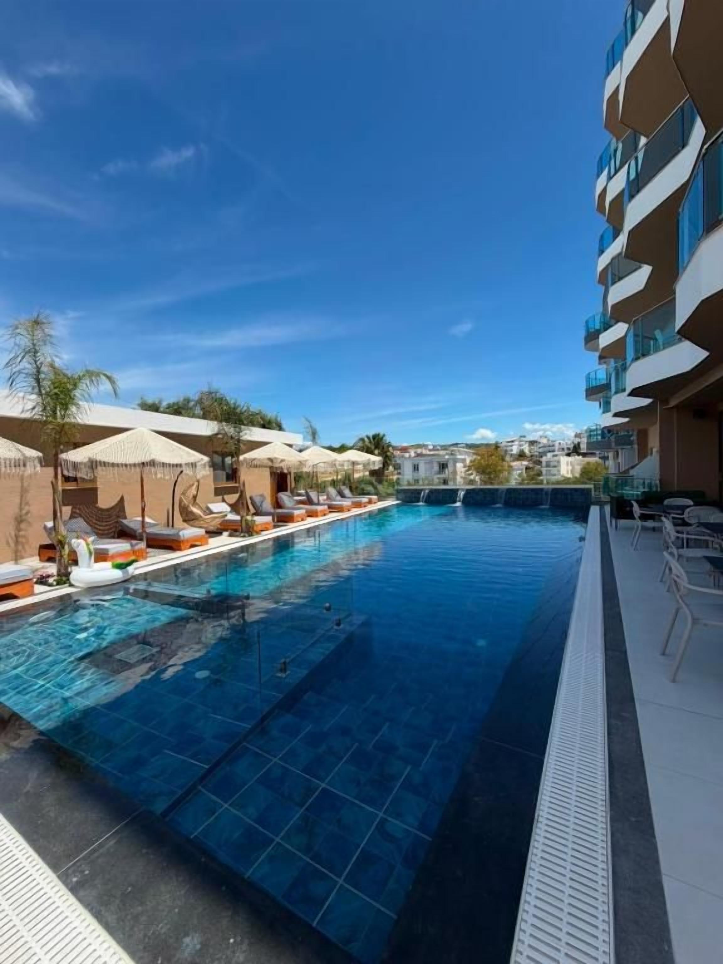 Una piscina con sillas de descanso y sombrillas, rodeada por un edificio con balcones.