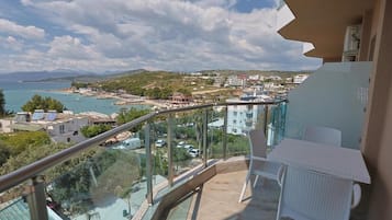Un balcón con baranda de vidrio que da a una ciudad costera, una mesa con sillas y una vista despejada del mar y edificios.