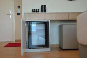 Un refrigerador compacto en una habitación con un armario de madera encima, una alfombra roja en el suelo y un sofá gris.