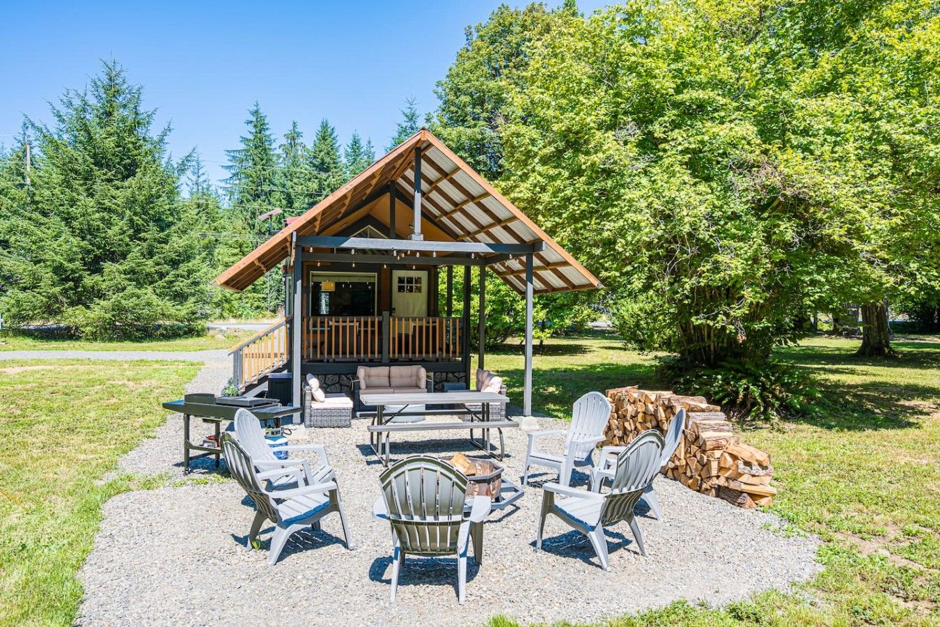 A covered outdoor seating area with a wooden deck, surrounded by chairs and a table, set against a backdrop of trees and a clear sky.