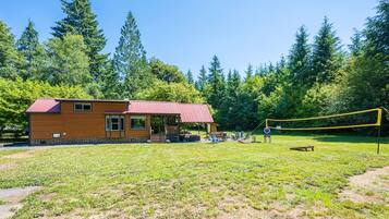 A wooden cabin with a red roof, a covered porch, and a volleyball net in a grassy area surrounded by trees.