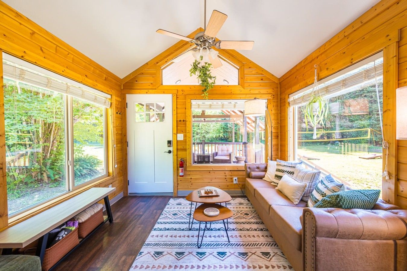 Interior acogedor de una cabaña con ventilador de techo, un sofá, una mesita y ventanas amplias que dan a vistas del exterior.