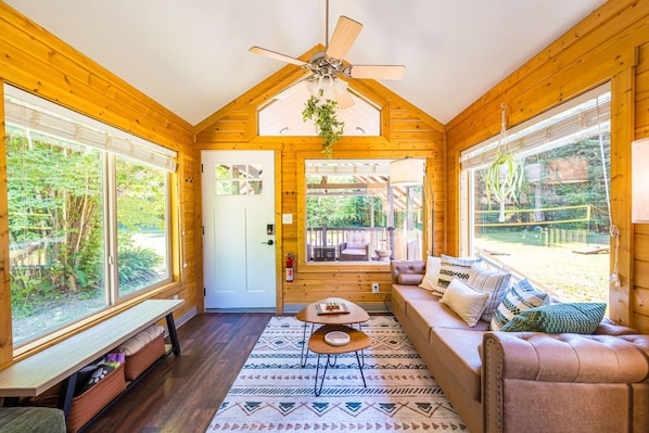 Interior acolhedor de uma cabana com ventilador de teto, um sofá, uma mesinha e grandes janelas com vista para o exterior.