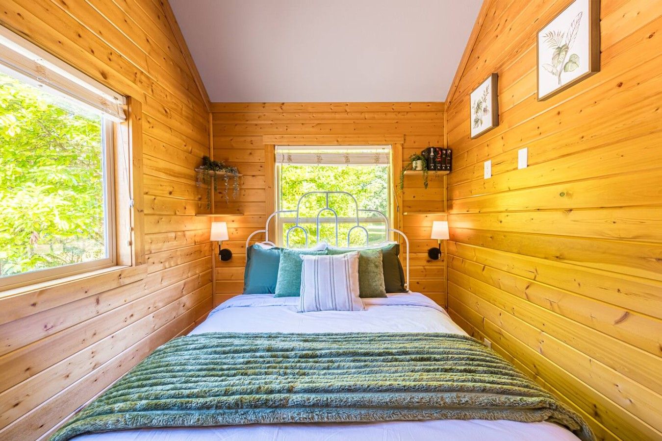 A wooden cabin bedroom with a bed, two wall-mounted lamps, and a window with a view of greenery.