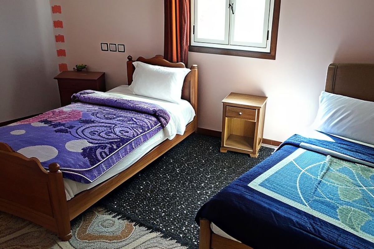 A bedroom with two beds, a nightstand, a small table, and a window.