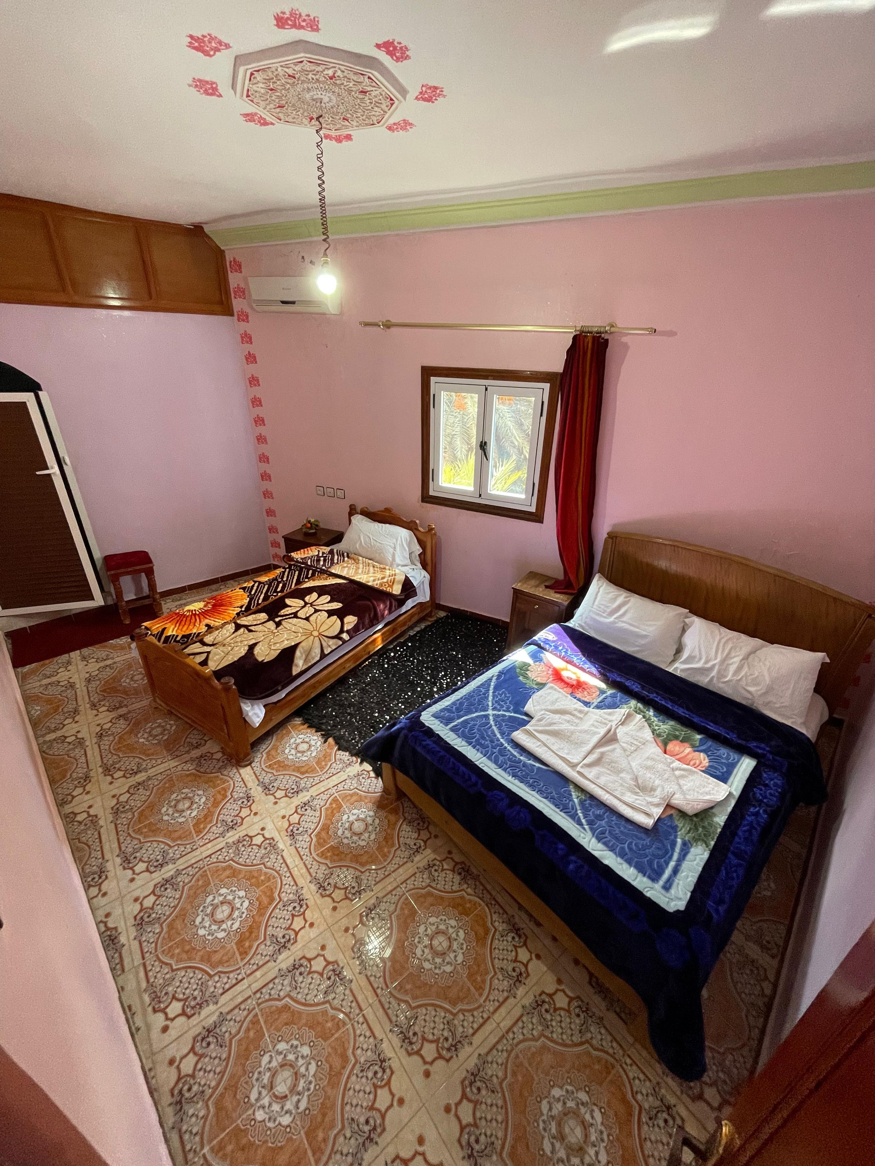 A bedroom with two beds, a window with curtains, a ceiling fan, and patterned floor tiles.