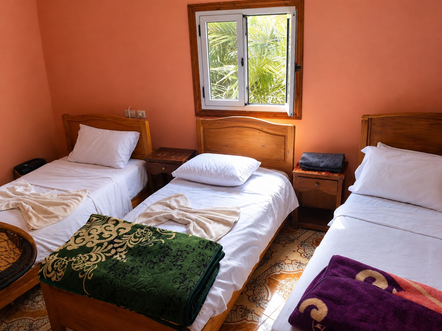 A room with two single beds, a window with a view of greenery, a wooden headboard, and a small bedside table with a lamp.