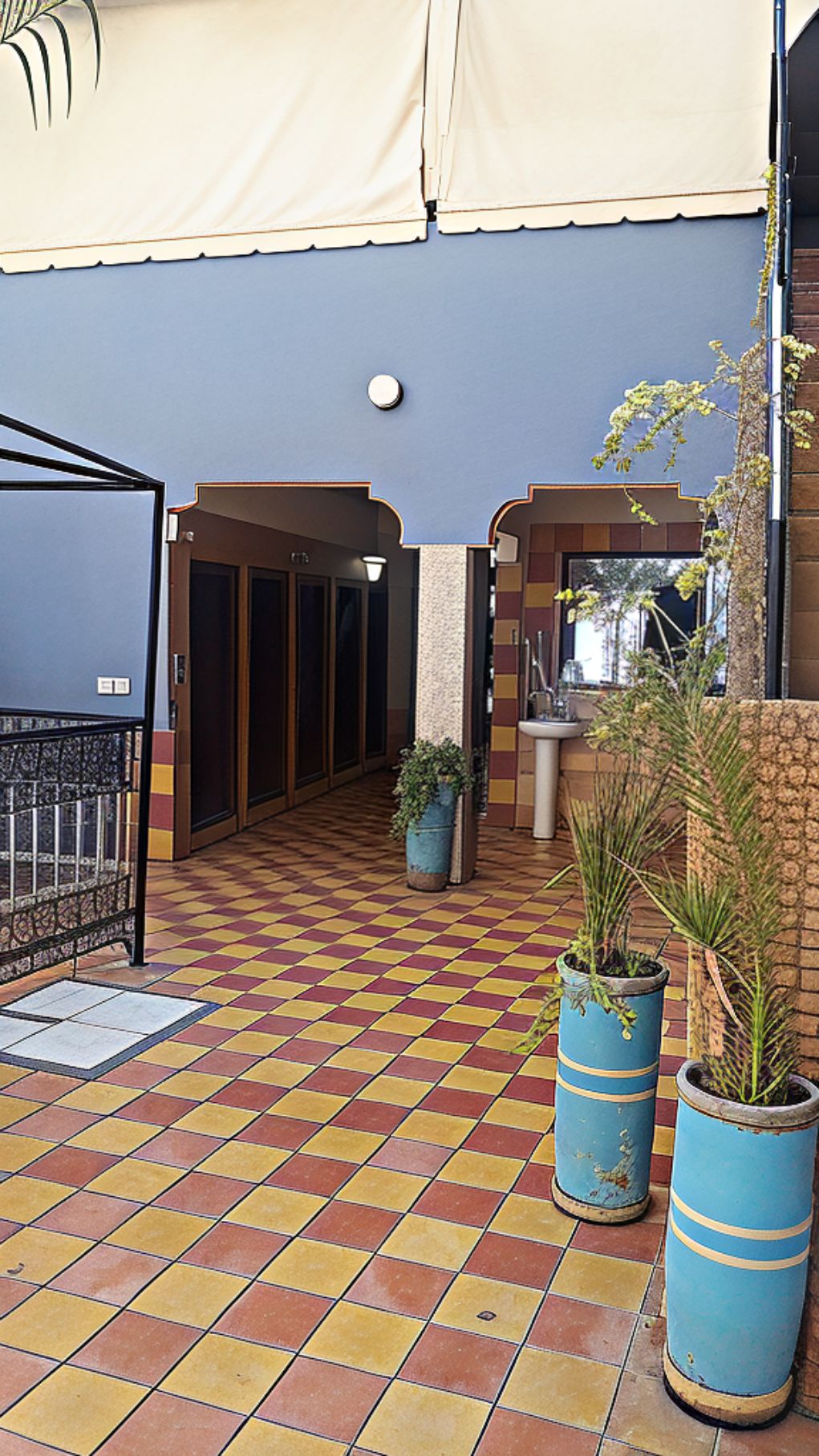 Corredor de hotel com piso xadrez, plantas em vasos e área coberta com luminária no teto.