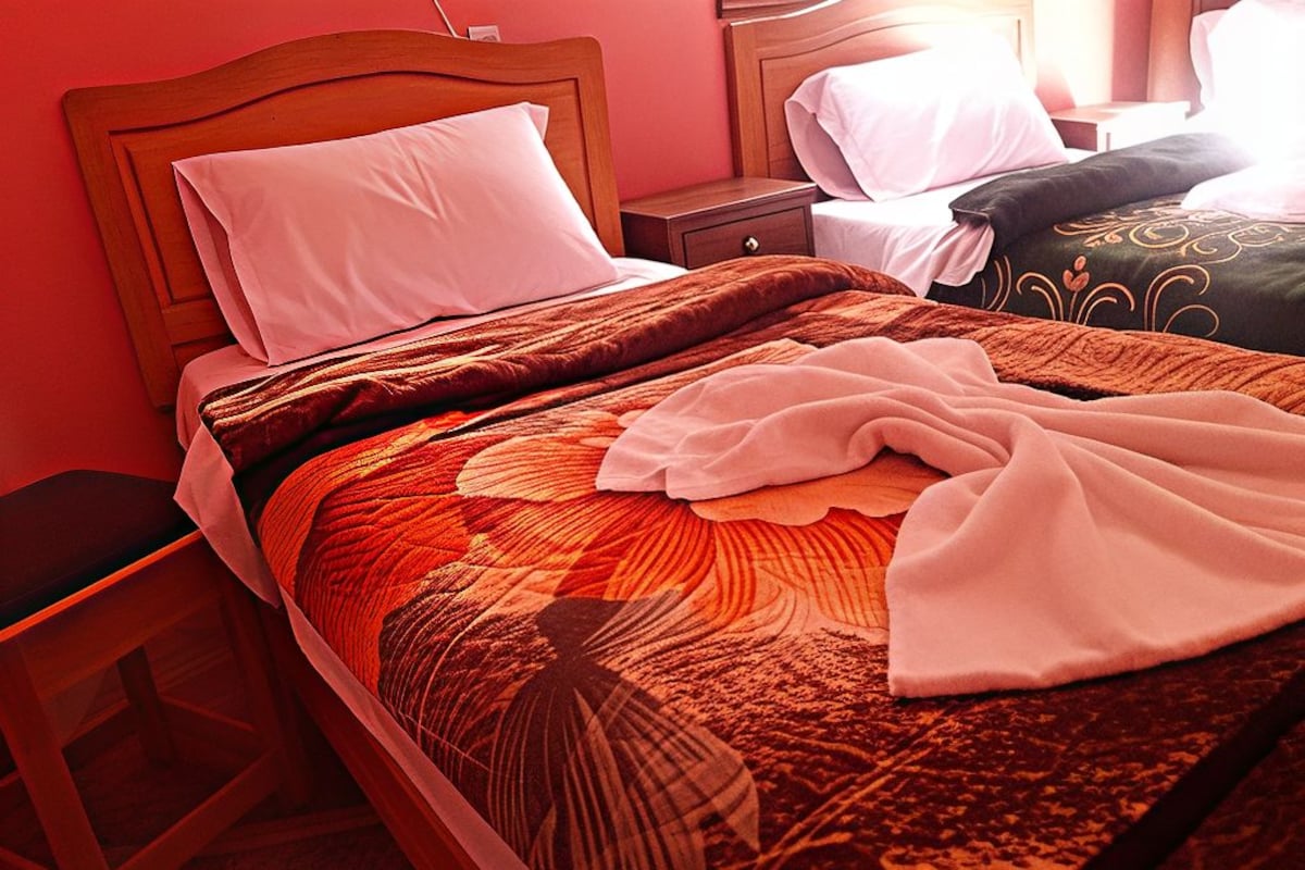 A hotel room with a wooden bed frame, a large bedspread with a leaf pattern, and a nightstand with a lamp.