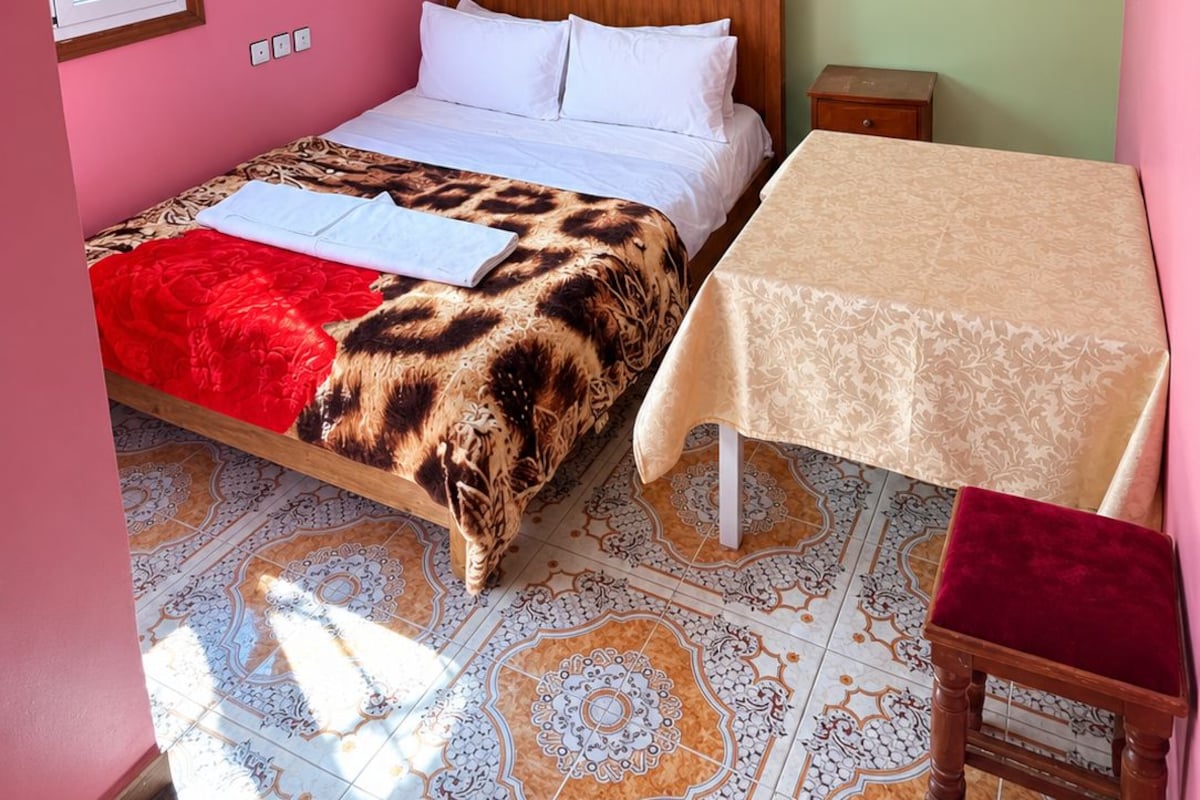 A room with a bed, a table, and a chair. There is a window with a view of greenery outside.