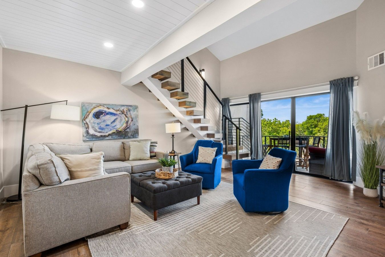 A spacious living room with a grey sofa, blue armchairs, a coffee table, and a staircase leading to a balcony with a view.