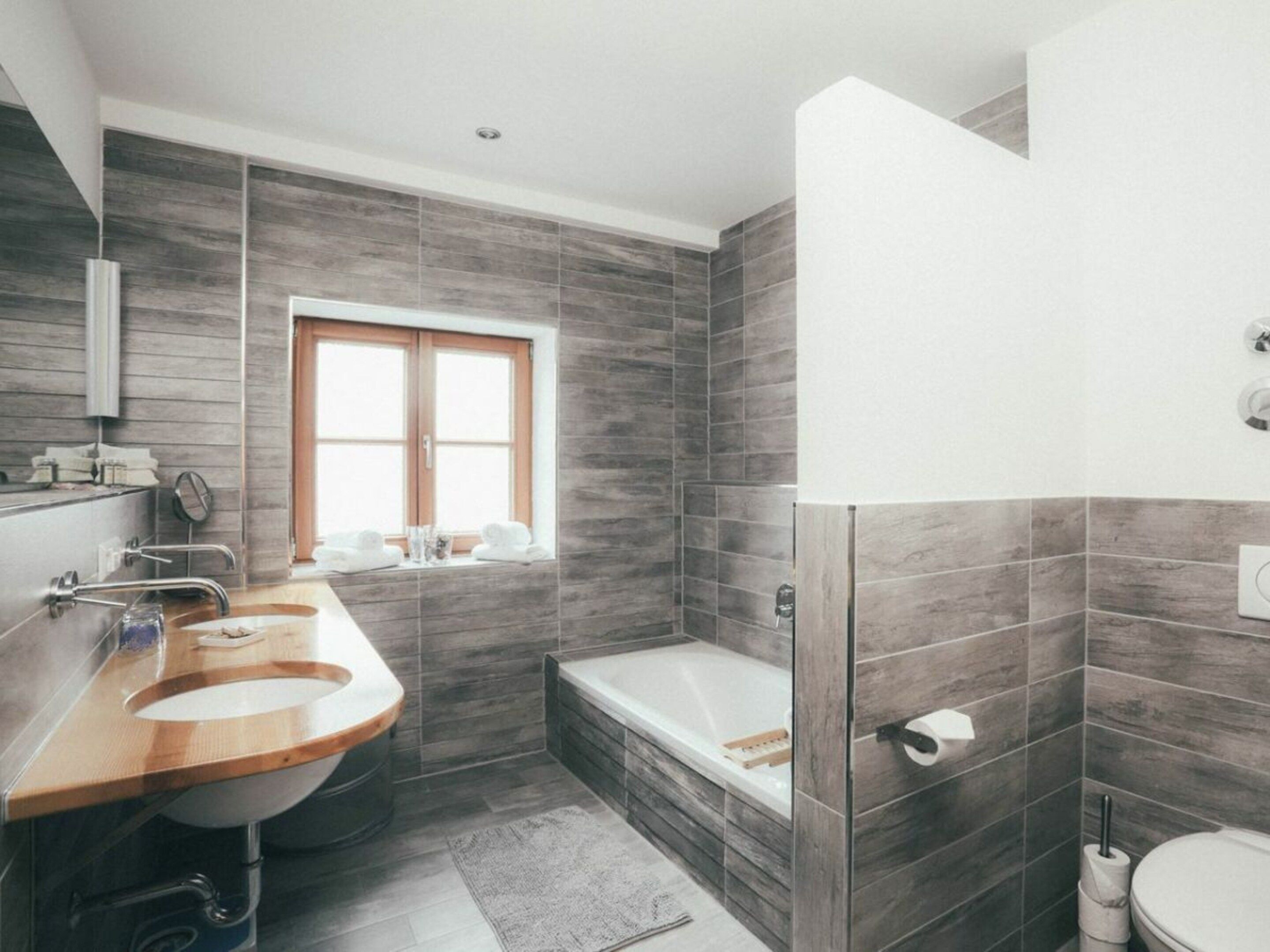 A modern bathroom with a wooden vanity, a bathtub, and a window.