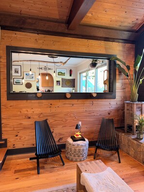 Une pièce aux murs en bois, avec un grand miroir, deux chaises noires, un pouf rond et une petite table sur laquelle se trouve une lampe.