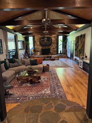 A spacious living room with a stone fireplace, a ceiling fan, and a large window. - Spiritual Retreat on 12-Acres in Travelers Rest - Sauna, Cold Plunge, Hottub (Travelers Rest)