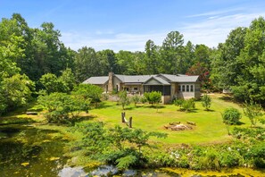 A house with a stone chimney, surrounded by a well-maintained lawn and a pond. - Spiritual Retreat on 12-Acres in Travelers Rest - Sauna, Cold Plunge, Hottub (Travelers Rest)