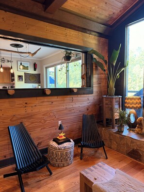 A cozy room with wooden walls, a large mirror, two black chairs, a small round ottoman, and a wooden coffee table. - Spiritual Retreat on 12-Acres in Travelers Rest - Sauna, Cold Plunge, Hottub (Travelers Rest)