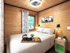 A cozy wooden room with a bed, a bedside table with a lamp, and a window with curtains. - Couples' Cabin 12 Min to Cherokee Casino | Pets (Whittier)