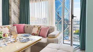 A modern living room with a sofa, a dining table set with plates and glasses, and a view of a cityscape through large windows.
