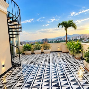 A rooftop terrace with a geometric patterned floor, potted plants, a spiral staircase, and a cityscape view in the background. - Hotel Signature StayZ Premium (Udaipur)