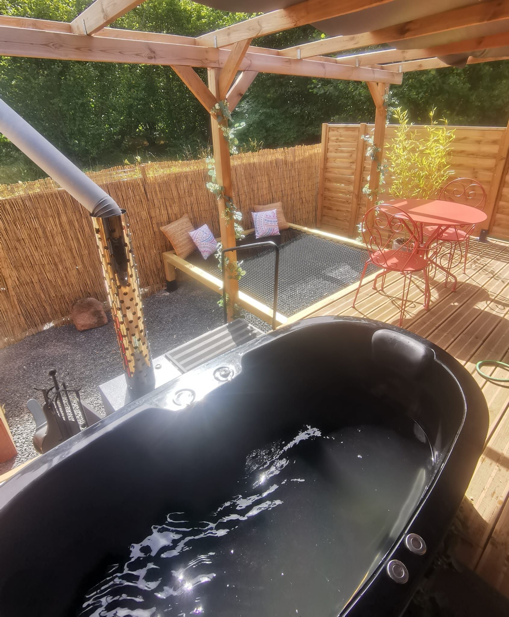 A hot tub in a wooden outdoor area with a pergola, a table with chairs, and a bamboo fence.