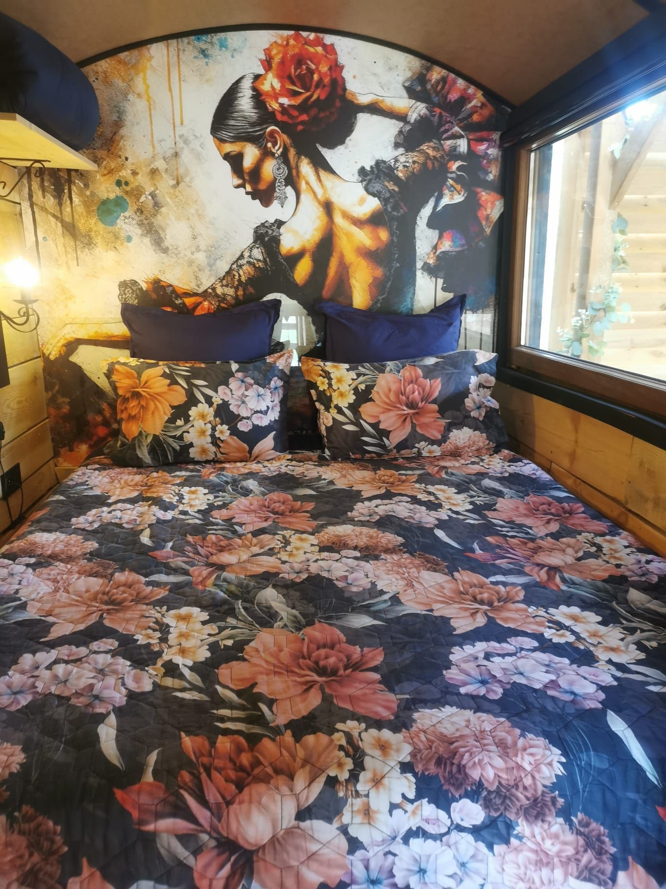 A bedroom with a floral bedspread, a large mural on the wall, and a window with a view.