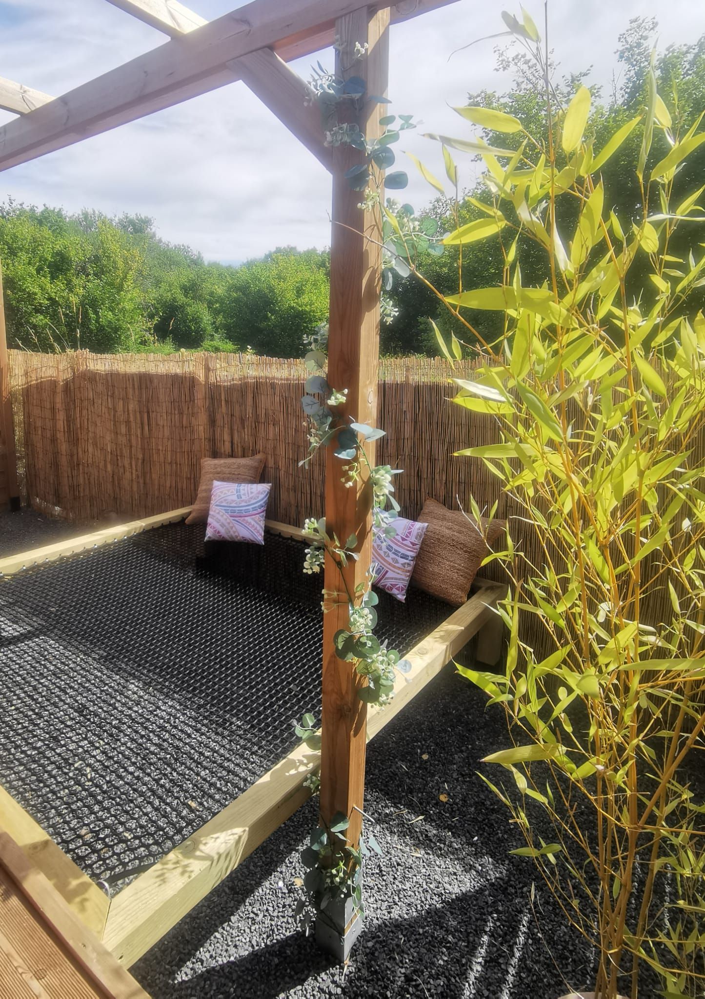 A wooden pergola structure with a woven bamboo wall, a black mesh floor, and two patterned cushions.