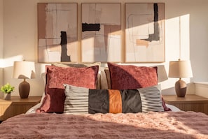 Une chambre à coucher avec un lit, deux tables de chevet, des lampes et des œuvres d’art abstraites accrochées au mur.