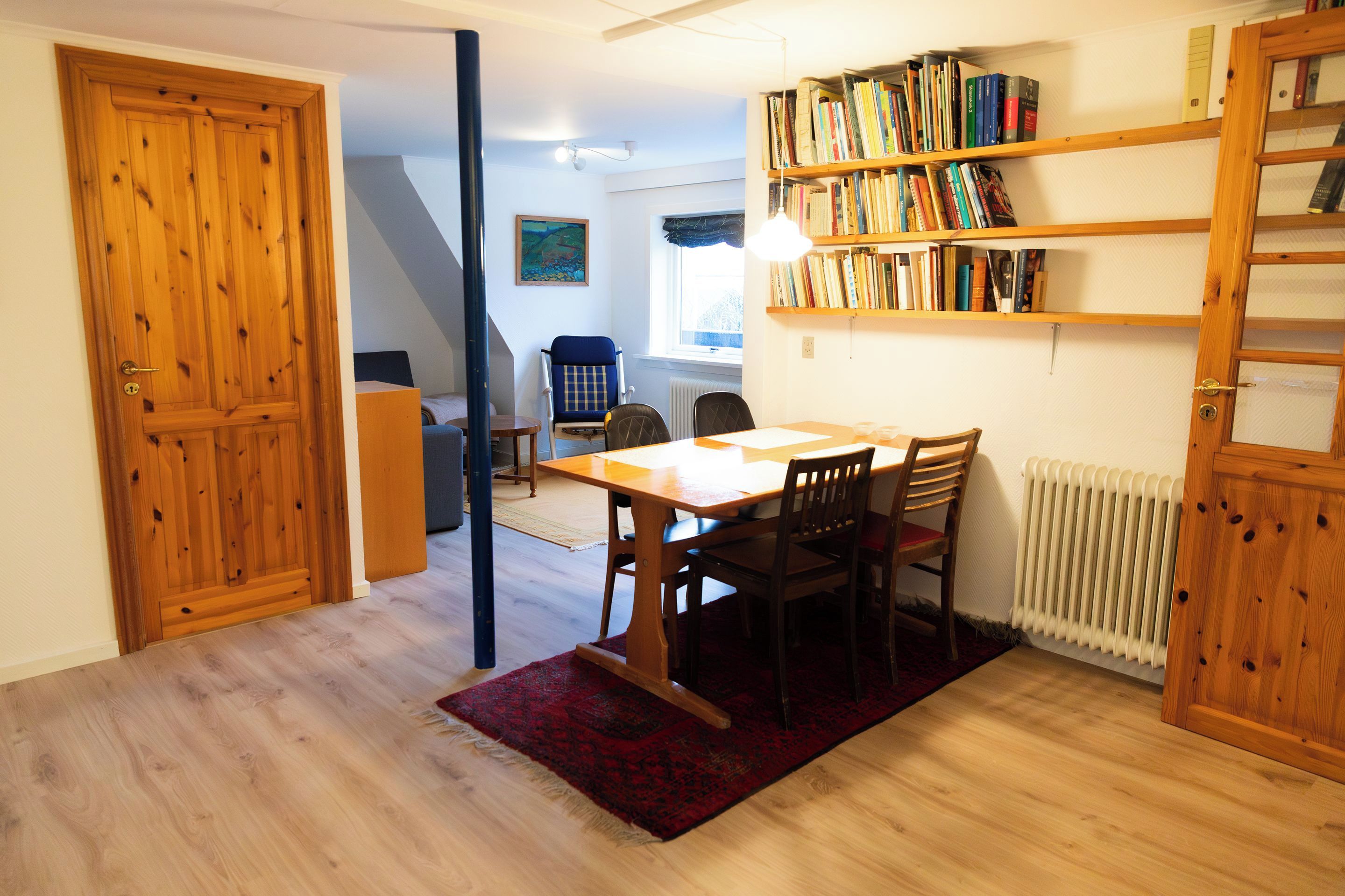 A dining area with a wooden table and chairs, a red rug, a bookshelf with books, and a radiator.