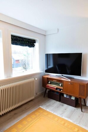 A room with a television on a wooden cabinet, a window with a black valance, and a radiator. - Hvarvið 9, ST. (Thorshavn)