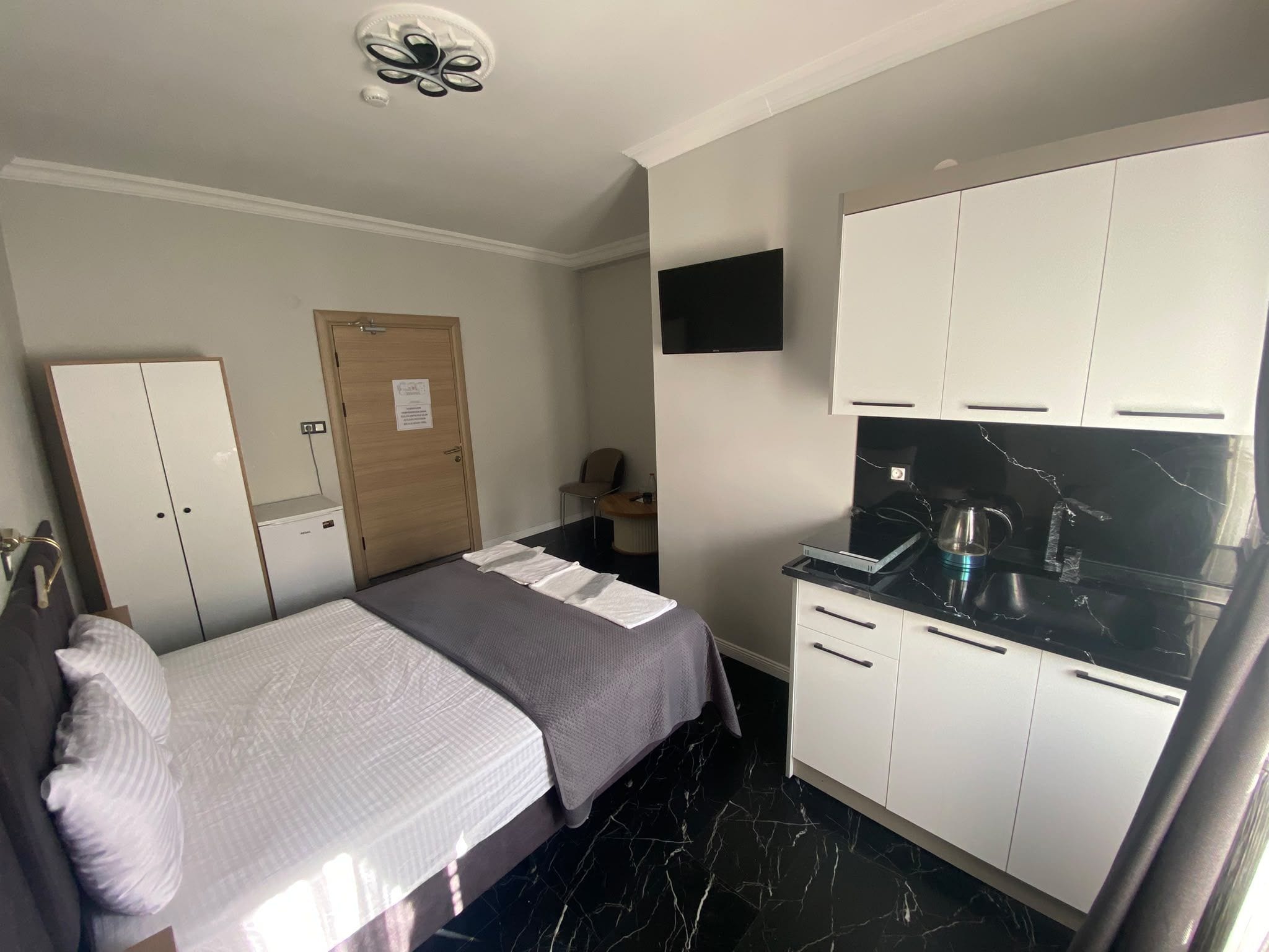 A compact room with a bed, a wardrobe, a small desk, a flat-screen TV, and a kitchenette with a stove.