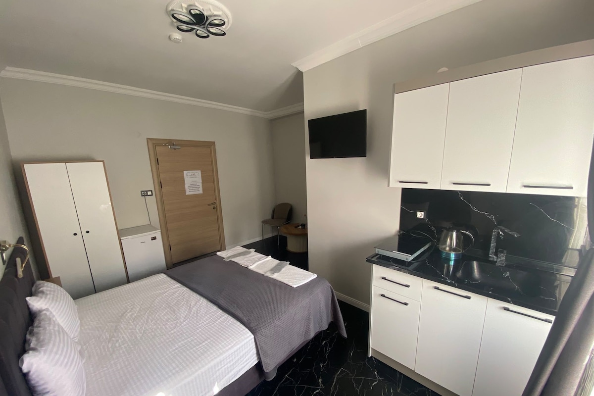 A compact room with a bed, a wardrobe, a small desk, a flat-screen TV, and a kitchenette with a stove.