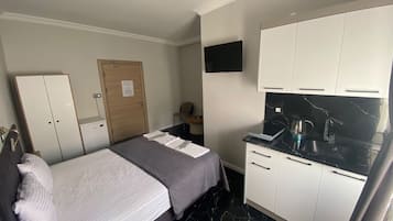 A compact room with a bed, a wardrobe, a small desk, a flat-screen TV, and a kitchenette with a stove.