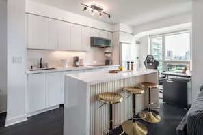 A modern kitchen with white cabinetry, a central island with stools, a built-in oven, and a large window offering a city view. - Lakefront Oasis Modern 1BR w Den (Toronto)