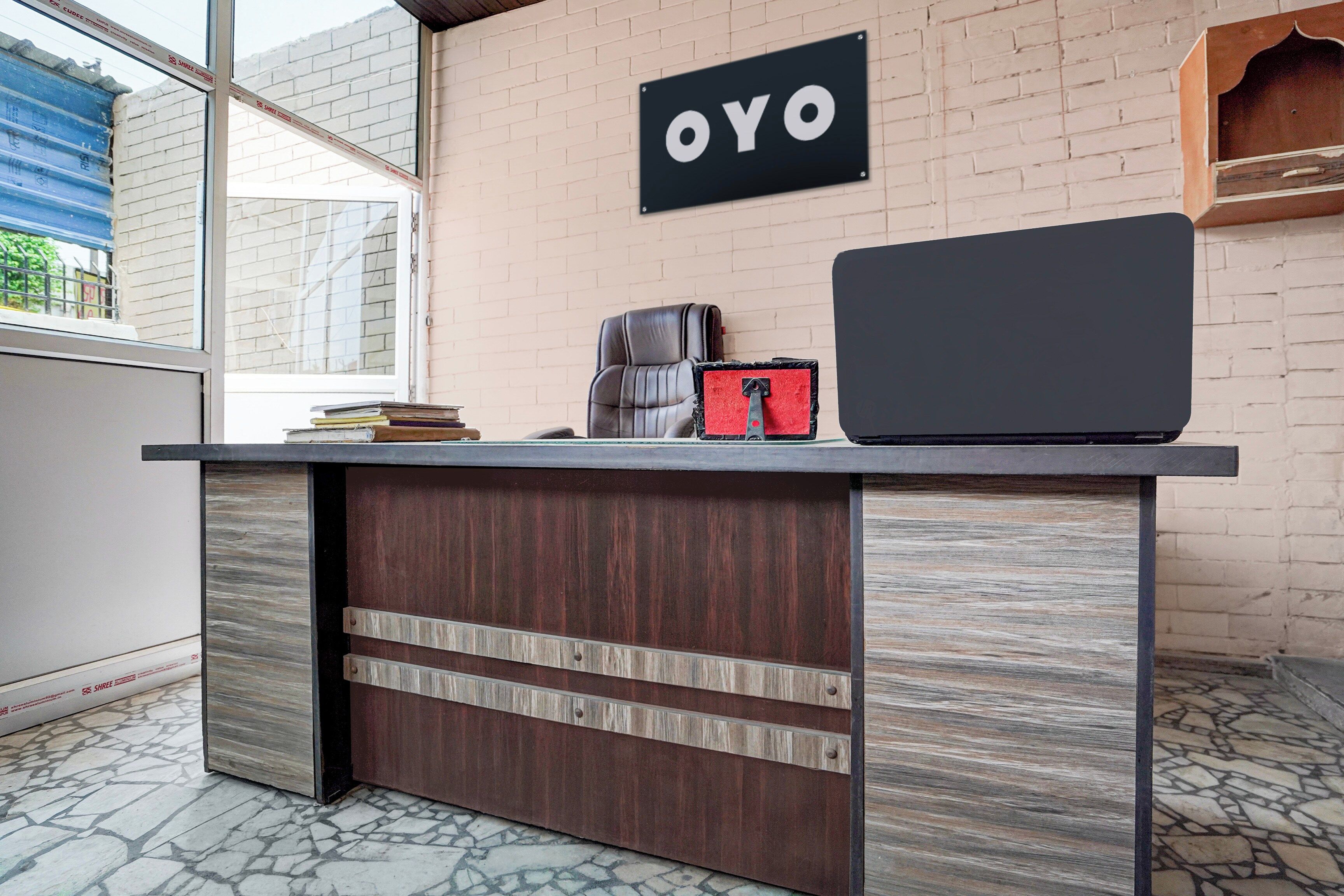 Bancada de recepção com um letreiro indicando 'OYO', uma cadeira de couro, um objeto vermelho e preto, e um laptop sobre o balcão.