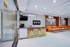 Ein Hotel-Empfangsbereich im modernen Design mit einer Rezeption, einer großen Wandtafel und einer Sitzecke mit orangefarbenen Sofas.