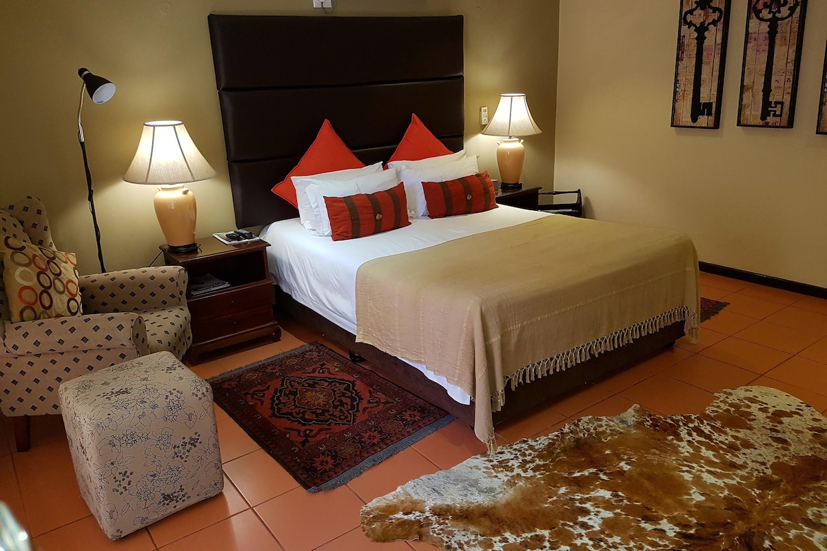 Una habitación de hotel con una cama, dos lámparas de noche, un mesita de noche, una silla, un tapete estampado y un tapete de cuero.