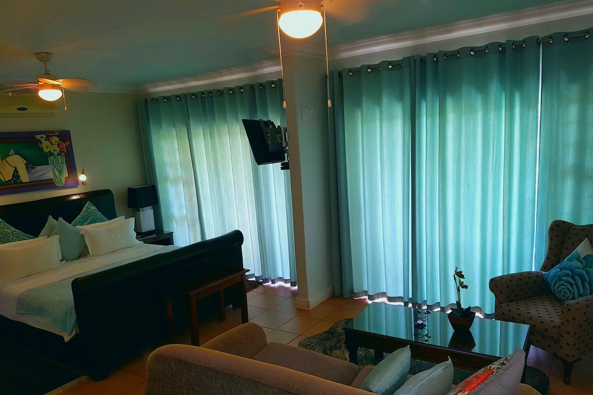 Una sala de estar con un sofá, un sillón y una mesa de centro. Hay un ventilador de techo y un televisor montado en la pared.