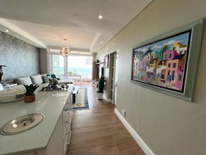 A modern kitchen with a central island, a dining area with a view, and a colorful painting on the wall. - Enjoyable 1-bedroom apartment in quaint Bloubergstrand neighborhood (Cape Town)