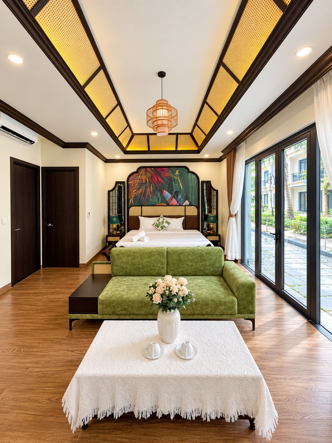 A hotel room with a large bed, a green sofa, a white table with a vase, and a view of the outside.