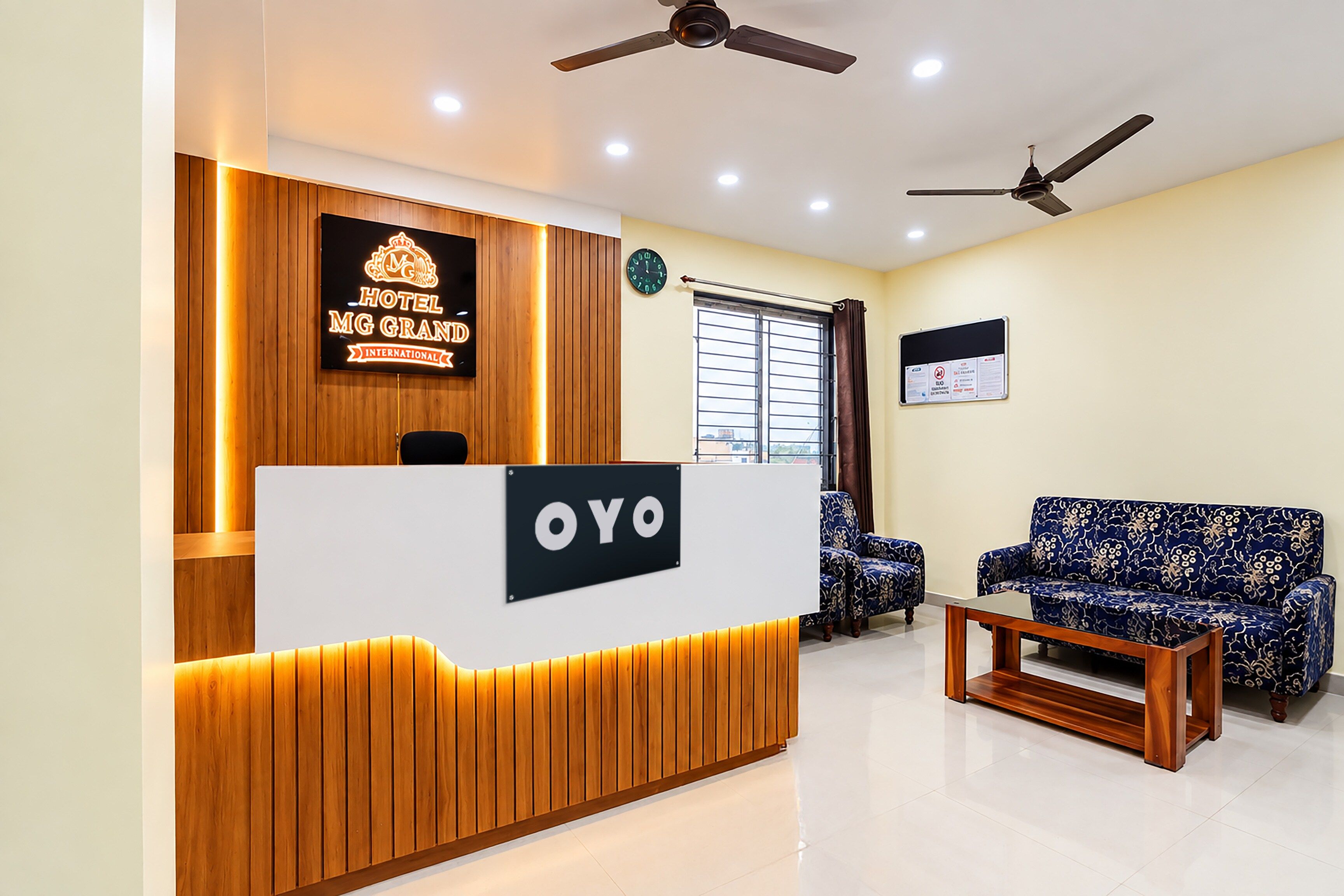 Un mostrador de recepción de hotel con un letrero que dice 'MG GRAND INTERNATIONAL' y un cartel encima que indica 'OYO'. Hay dos ventiladores de techo, un reloj y una ventana con cortinas.
