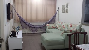 A room with a sofa, a small table, a chair, a hammock, and a window with curtains. - New Listing (Guarujá)
