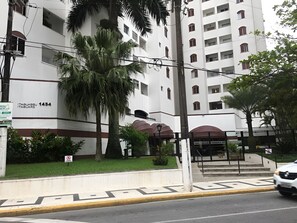 A multi-story building with a flat facade, numbered 1454, featuring a central entrance with a covered area and a staircase leading to the entrance. - New Listing (Guarujá)