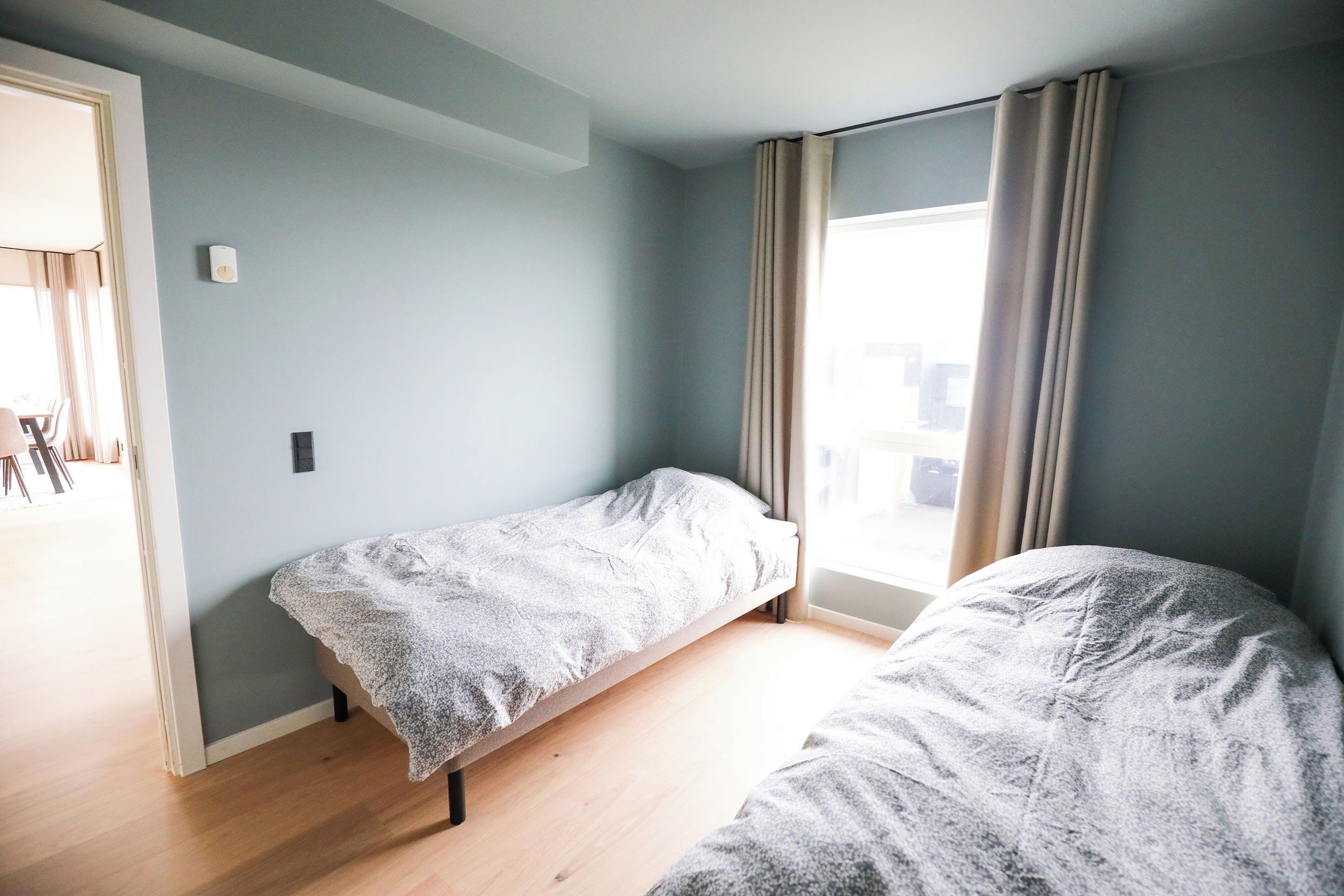 Una habitación con dos camas individuales, una ventana con cortinas, una puerta que da a otra habitación y un interruptor de luz en la pared.