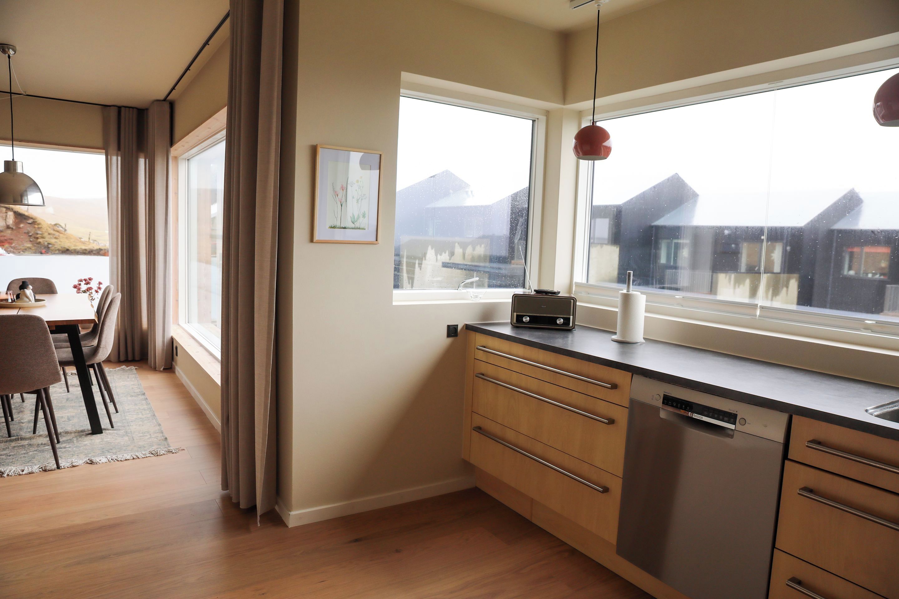 A modern kitchen with a dishwasher, a window with a view of buildings, and a dining area with a table and chairs.
