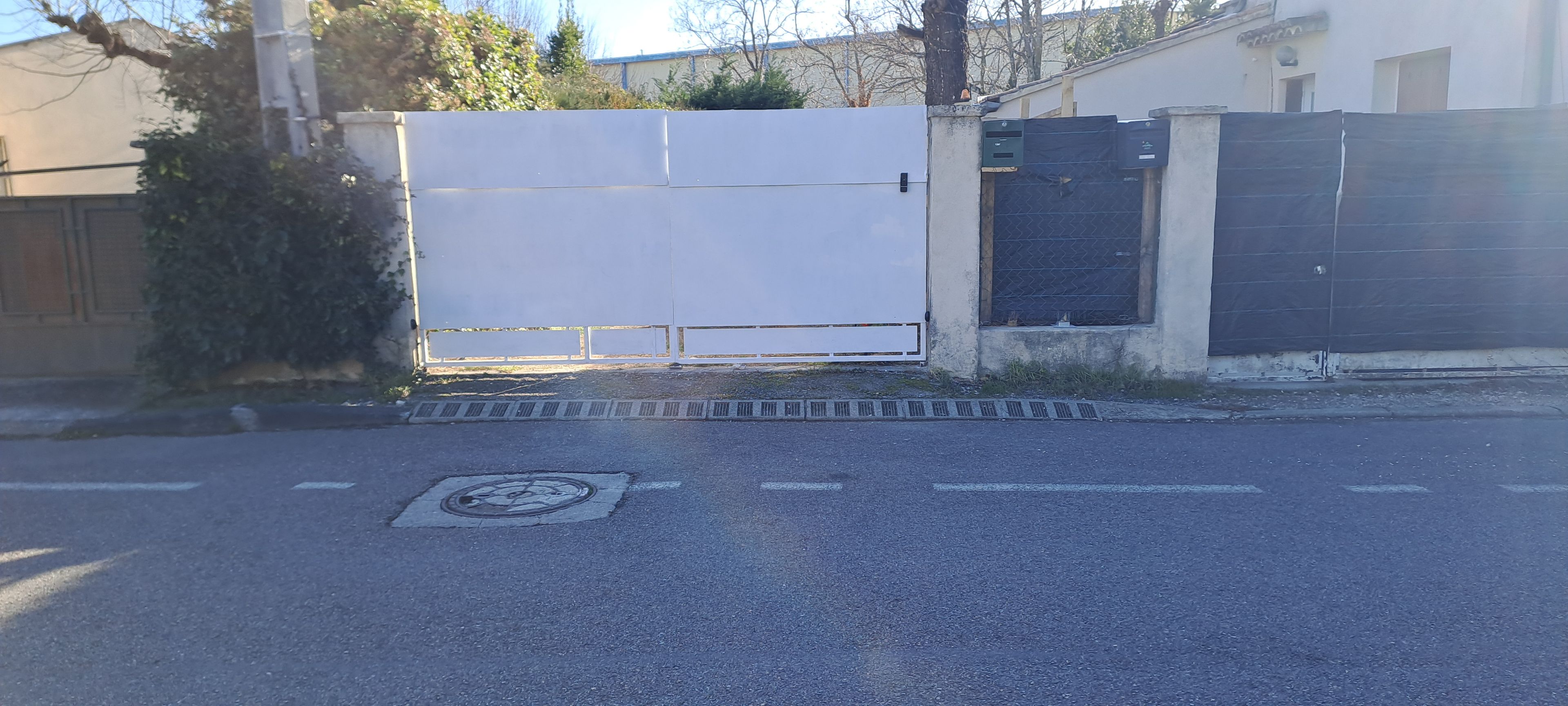 Een toegangshek met een metalen poort, een betonnen pad en een putdeksel in de weg.