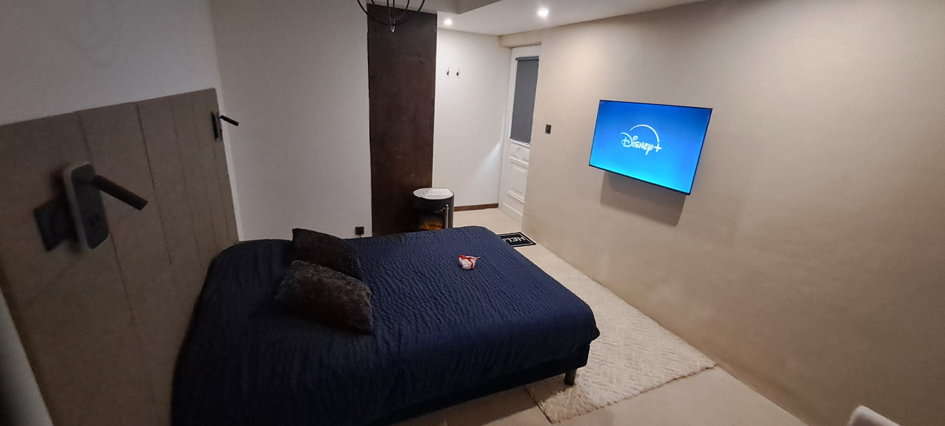 Een slaapkamer met een bed, een aan de muur bevestigde televisie waarop het Disney+-logo te zien is, en een klein nachtkastje.