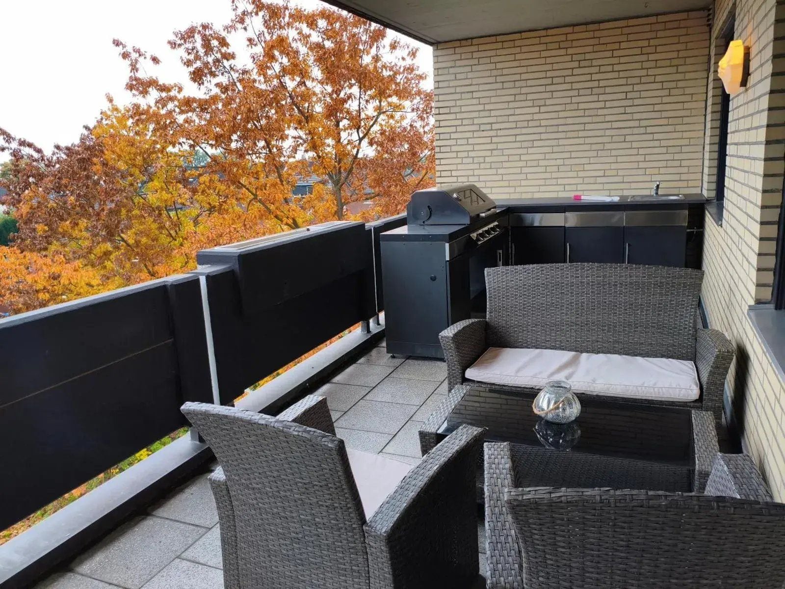 Ein Balkon mit Korbmöbeln, einem Grill und Blick auf herbstliche Bäume.