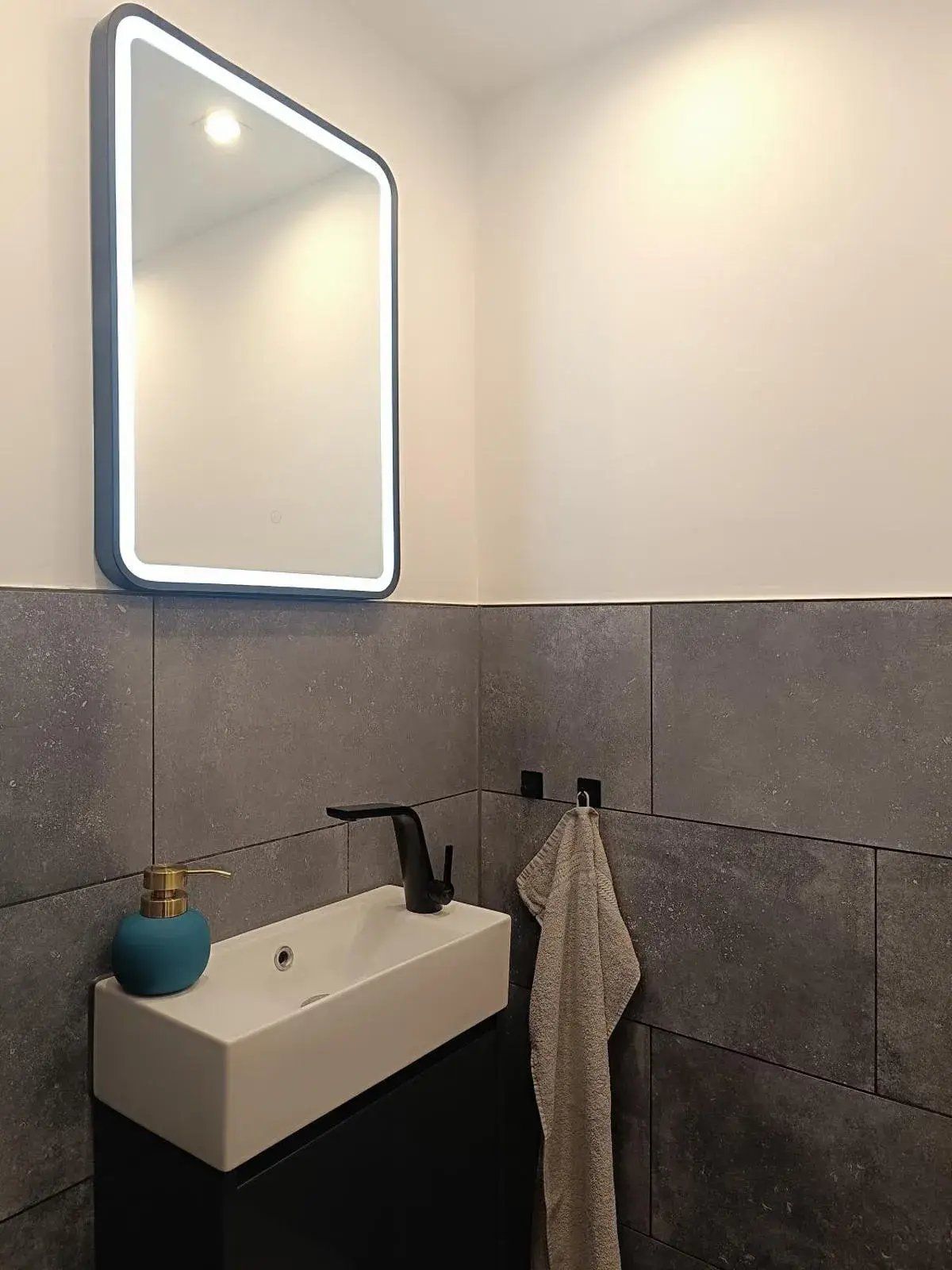 Ein modernes Badezimmer mit einem rechteckigen Spiegel, einem weißen Waschbecken, einem schwarzen Wasserhahn, einem blauen Seifenspender und einem Handtuch an einem Haken.